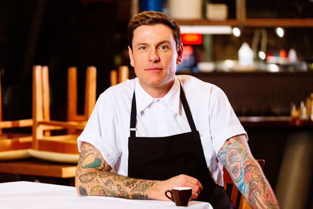 Headshot of Chef Chuck Hughes in a darkened restaurant, wearing a black apron over a white button-up shirt.