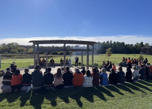 A group of people sitting in a circle around the A’nó:wara Learning Circle with a firepit in the middle for a ceremony.