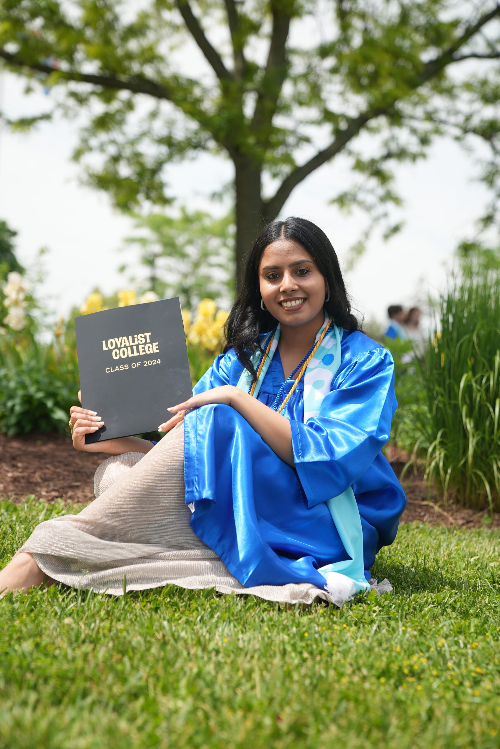 Female graduate in a blue gown and Loyalist Alumni scarf sitting on grass, holding a black diploma folder that says 'Loyalist College Class of 2024,' with trees and flowers in the background.