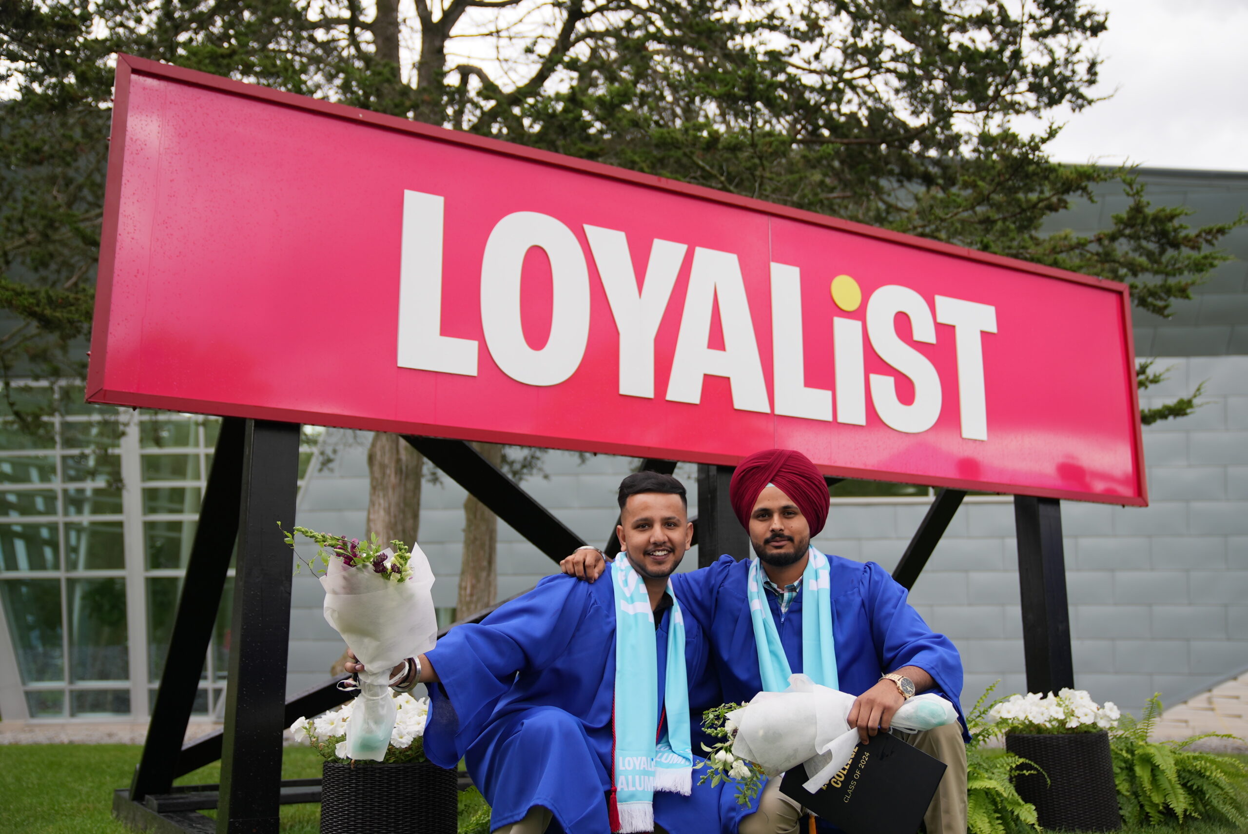 Two male graduates in blue gowns and Loyalist Alumni scarves crouch together in front of a large red 'Loyalist' sign, both holding bouquets of flowers and smiling at the camera.