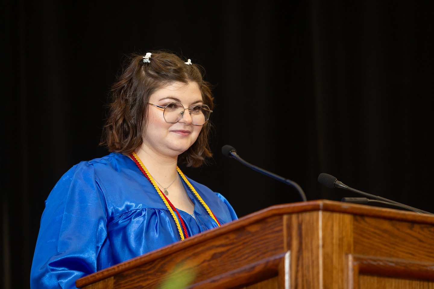 Young woman wearing a blue graduation gown and cords, standing at a wooden podium and speaking into a microphone. She has short hair with small white clips and is wearing glasses. The background is dark.