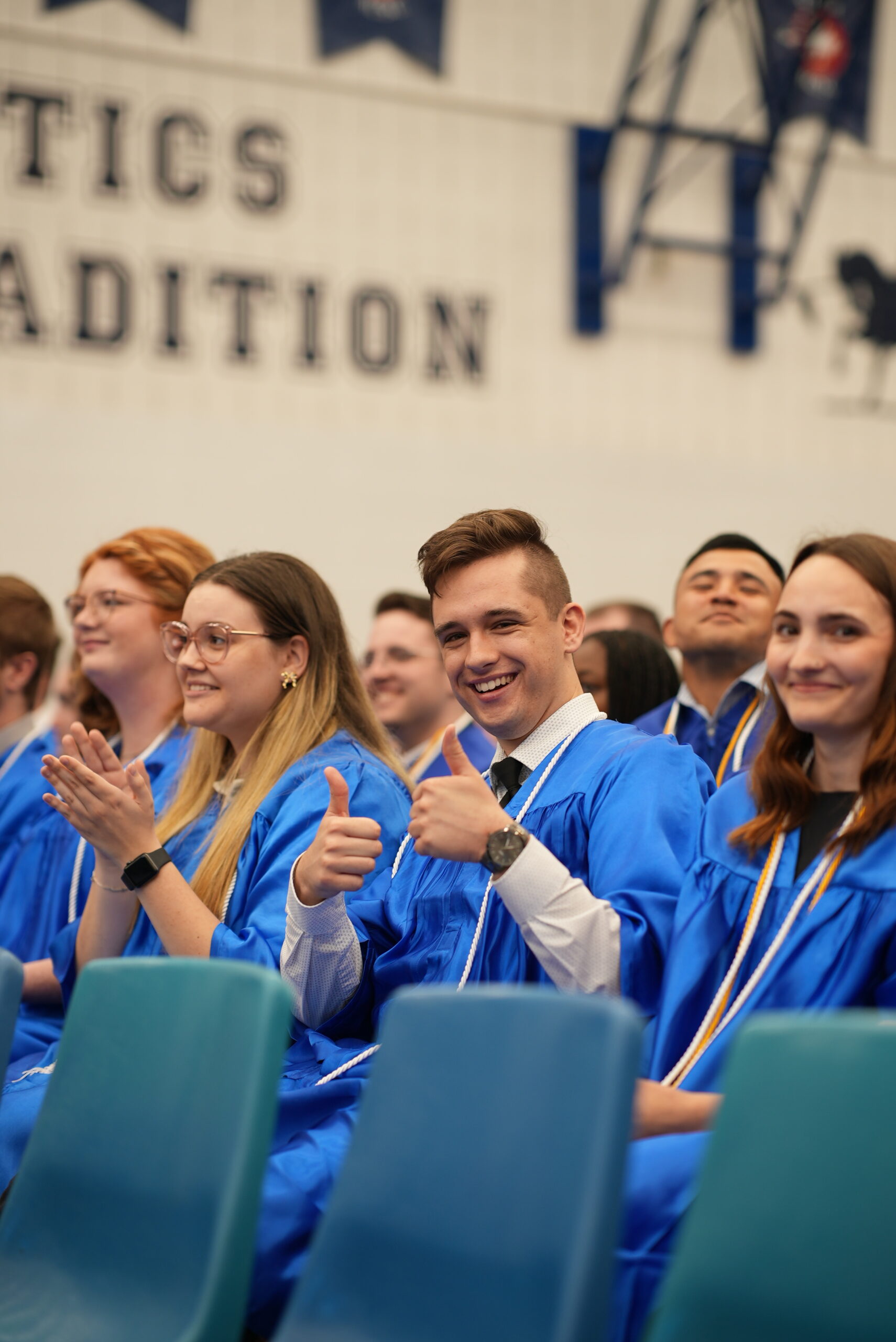 A group of graduates in blue gowns sitting in chairs during a ceremony. A young man in the centre smiles and gives two thumbs up, while others around him clap and smile. The setting is inside the gym at Loyalist's Belleville location.