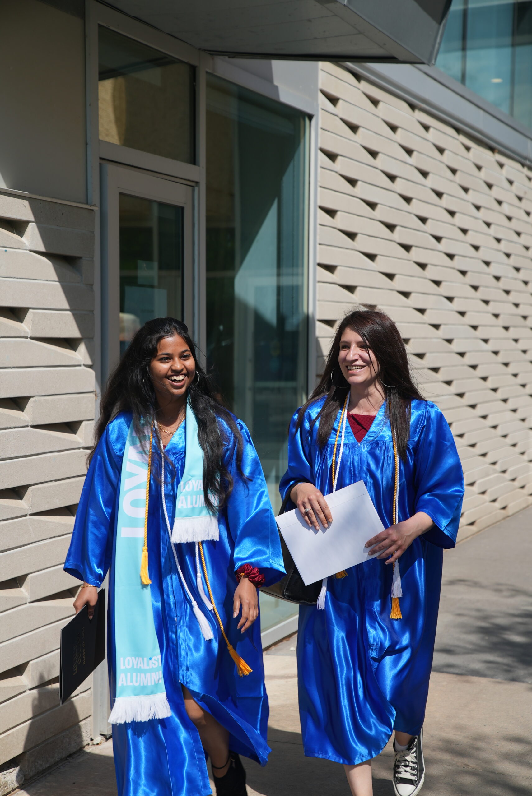 Two female graduates in blue gowns smiling as they walk outside the Parrot Centre of the Kente Building at the Belleville location. One holds a diploma folder, and the other has a Loyalist Alumni scarf.