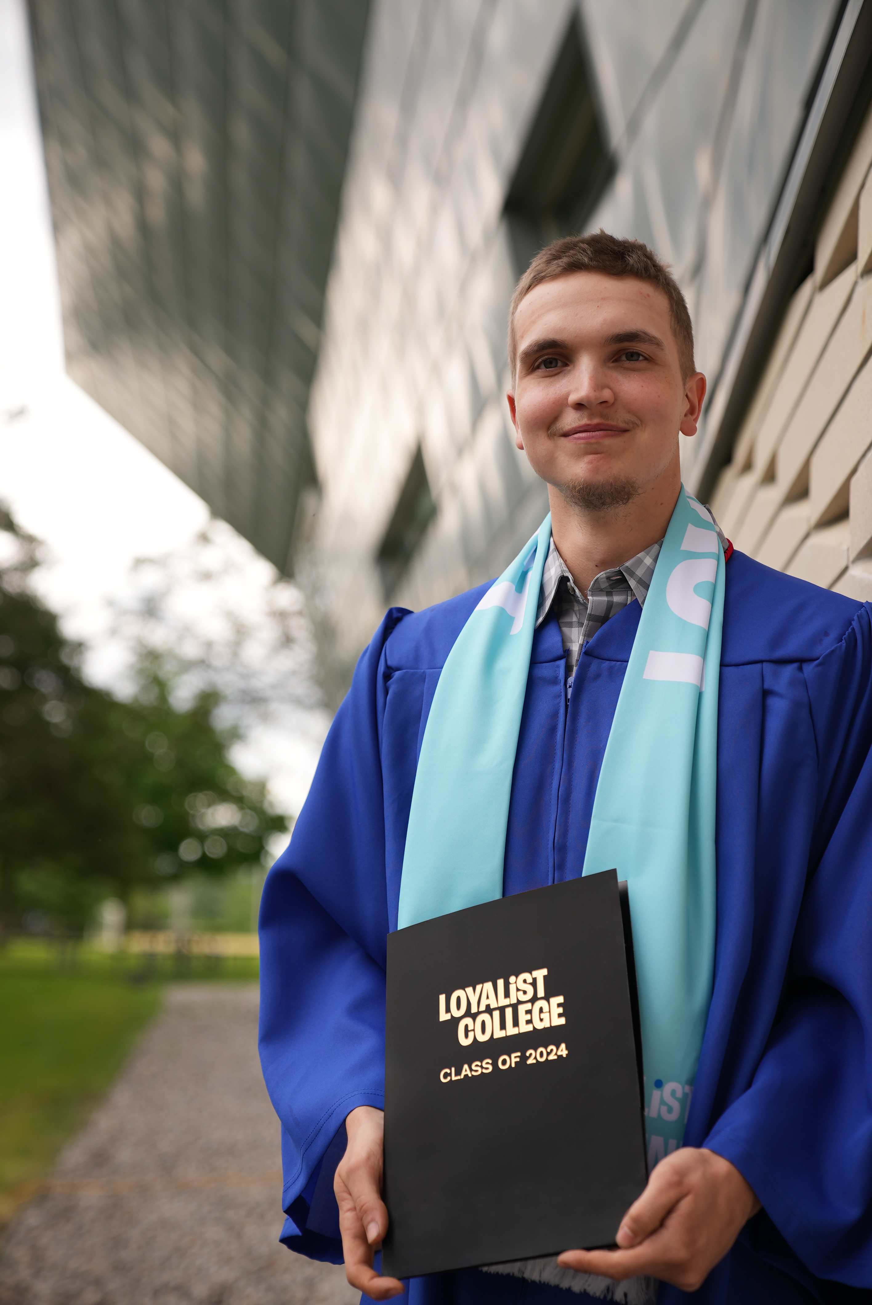 Male graduate in a blue gown and Loyalist Alumni scarf holding a black diploma folder that says 'Loyalist College Class of 2024' while standing outdoors near the Parrot Centre at the Kente Building at the Belleville location.