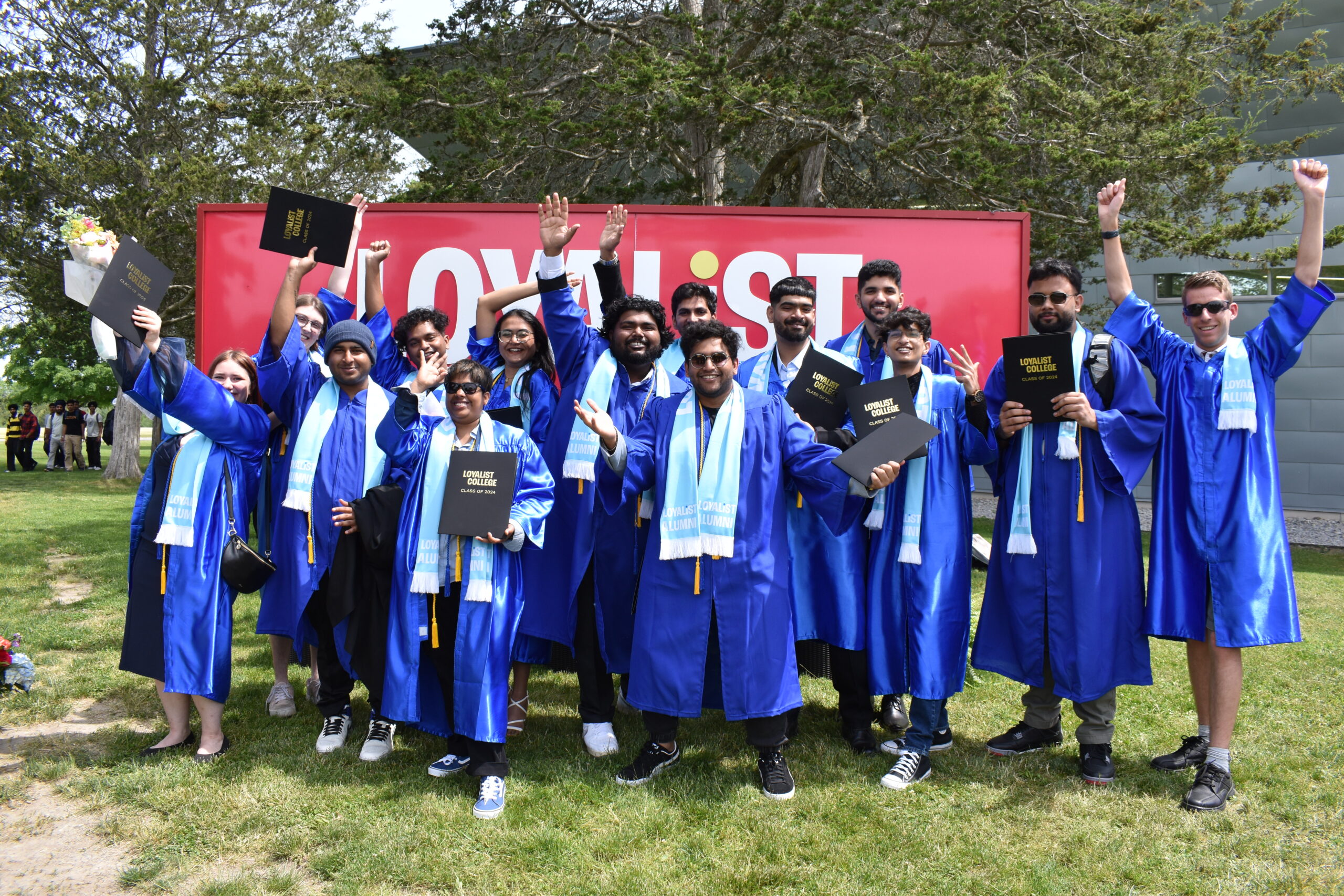 A group of graduates wearing blue gowns and alumni scarves celebrating outdoors. They are holding their diplomas and raising their hands in excitement. The backdrop features a large red 'Loyalist' sign, and trees are visible in the background.