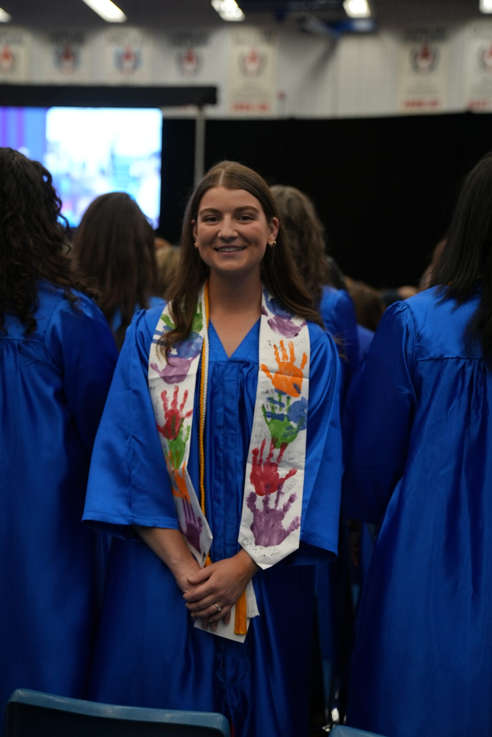 Smiling female graduate in a blue gown standing during a ceremony, wearing a colourful sash decorated with handprints. The background shows other graduates also in blue gowns, standing and facing the stage.