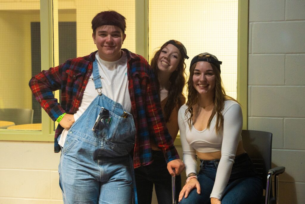 Three students dressed in casual, country-themed clothes smile for the camera during a Country Night event. One person wears denim overalls and a flannel shirt, while the other two wear jeans, crop tops, and backward baseball caps. They are sitting and standing in the Shark Tank Pub at Loyalist College.