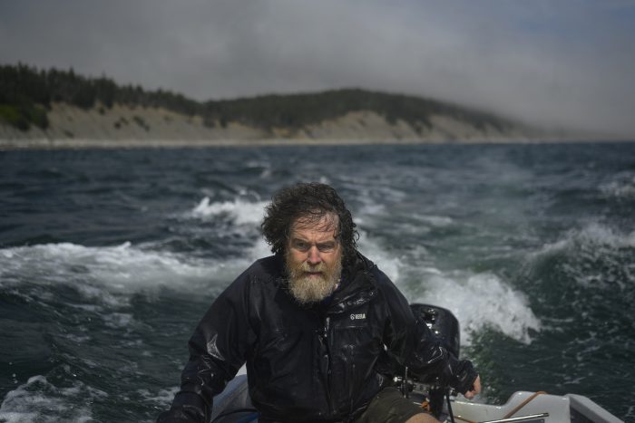 Man with a wet, scruffy beard and windblown hair, wearing a rain jacket, driving a small boat on choppy waters. The background shows a misty shoreline with trees and hills in the distance under an overcast sky.