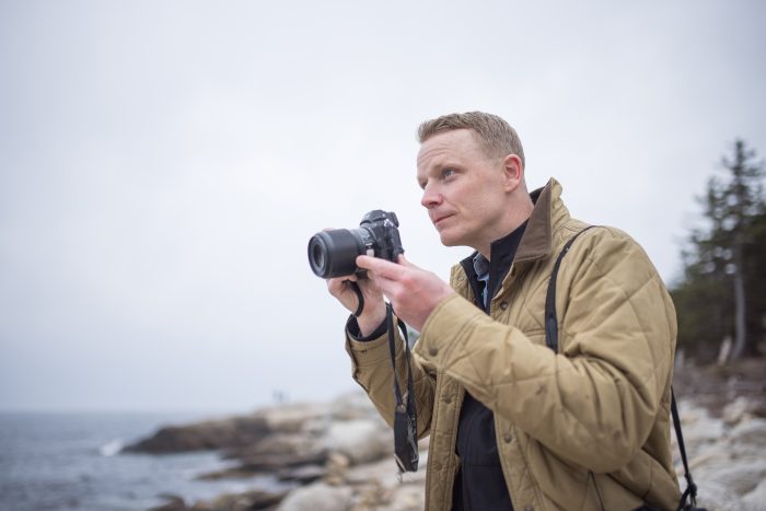 Loyalist College Photojournalism graduate Darren Calabrese, looking away and holding a camera pointed out of frame. He's wearing a brown jacket, and in the background is a rocky shoreline.