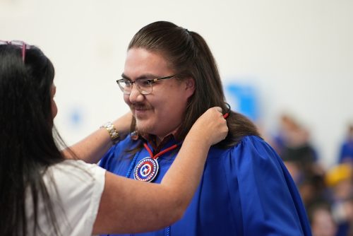 A person places a beaded medallion around a student's neck. The student is wearing a blue graduation gown.