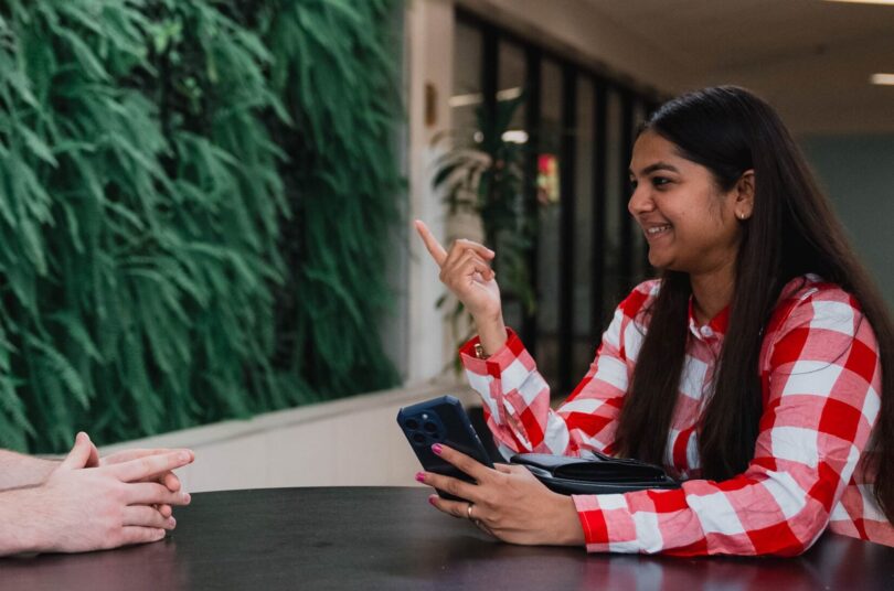 Two students sit at a table in front of a green living wall. One student holds a mobile phone and is using her hands to gesture while speaking.