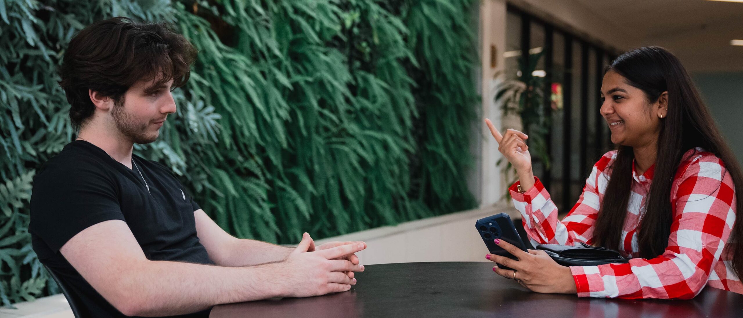 Two students sit at a table in front of a green living wall. One student holds a mobile phone and is using her hands to gesture while speaking.