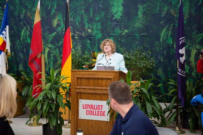 Dr. Ann Marie Vaughan, President and CEO, at a podium in front of a fake plant backdrop, speaking to an audience not shown.