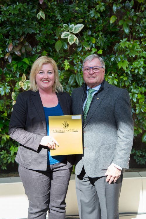 Ashley Cooney, a Loyalist Business student presented with an award by Ken Wheeler from the Belleville Rotary Club. They're standing in front of a live greenery wall in the Loyalist Link Lounge.