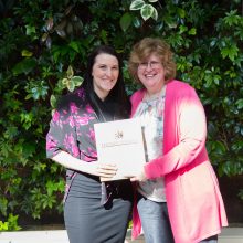 Darcy Eligh, a Loyalist Public Relations student, is presented with an award by Dr. Ann Marie Vaughan, President and CEO. They're standing in front of a live greenery wall in the Loyalist Link Lounge.