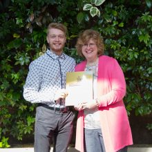 Josh Roberts, a Loyalist Public Relations student, is presented with an award by Dr. Ann Marie Vaughan, President and CEO. They're standing in front of a live greenery wall in the Loyalist Link Lounge.