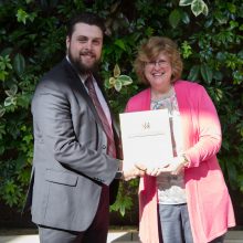 Scott Rook, a Loyalist Public Relations student, is presented with an award by Dr. Ann Marie Vaughan, President and CEO. They're standing in front of a live greenery wall in the Loyalist Link Lounge.