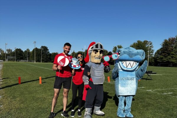 Two members of the student government stand on a grassy field, holding fun beach-themed props. They are accompanied by two mascots: a knight in silver armor and a shark wearing a Loyalist College jersey. The group poses for the camera under a bright, clear sky, enjoying a sunny day outdoors.