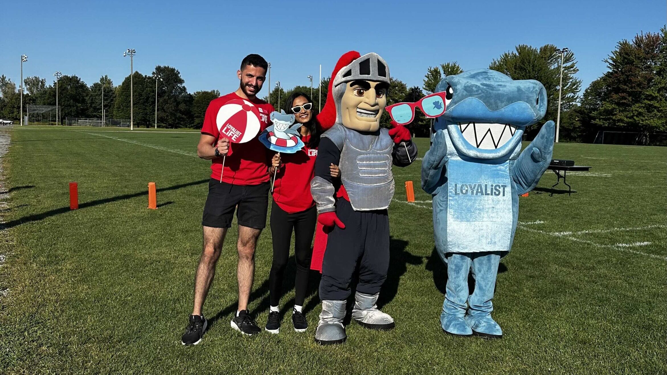 Two members of the student government stand on a grassy field, holding fun beach-themed props. They are accompanied by two mascots: a knight in silver armor and a shark wearing a Loyalist College jersey. The group poses for the camera under a bright, clear sky, enjoying a sunny day outdoors.