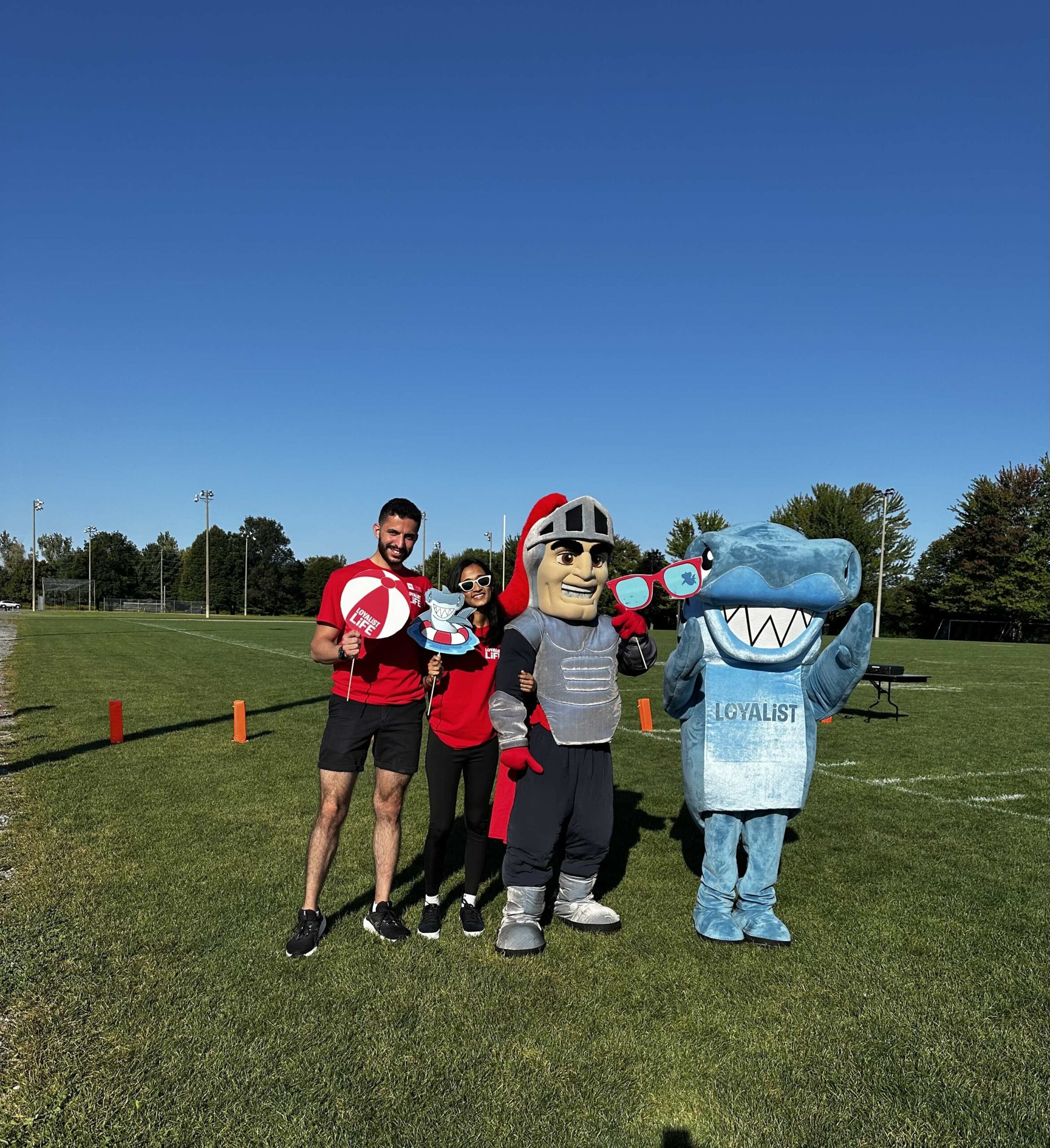 Two members of the student government stand on a grassy field, holding fun beach-themed props. They are accompanied by two mascots: a knight in silver armor and a shark wearing a Loyalist College jersey. The group poses for the camera under a bright, clear sky, enjoying a sunny day outdoors.