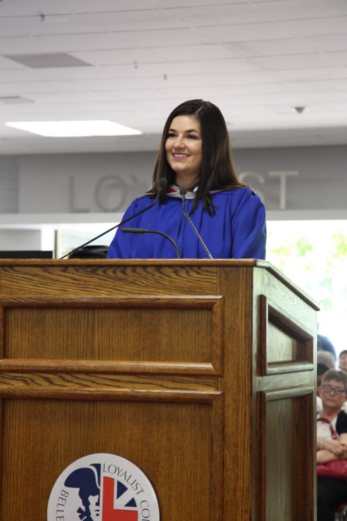 Graduate Natasha Mathieu, valedictorian, standing at a Loyalist College podium in a blue graduation gown, addressing an unseen audience.