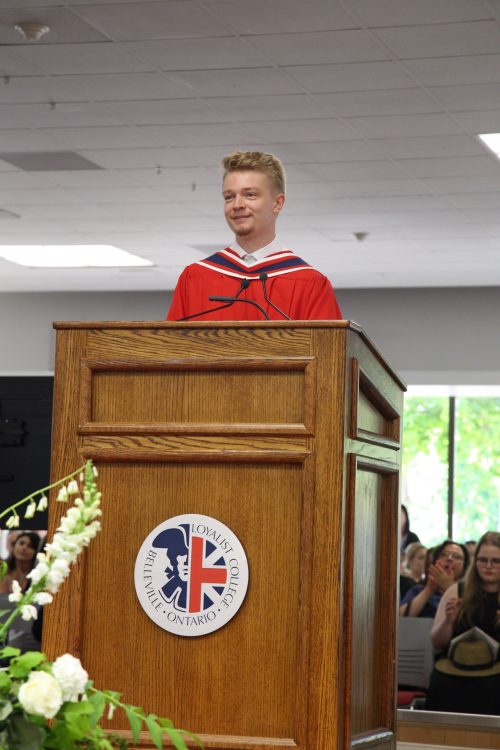 Graduate Josh Roberts, valedictorian, standing at a Loyalist College podium in a red graduation gown, addressing an unseen audience.