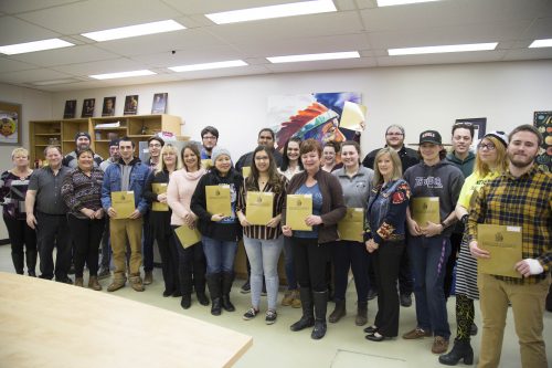 A group of students, standing in a classroom holding certificates. The group is arranged in two rows, with some smiling directly at the camera. The room has bright fluorescent lighting, and shelves with books and artwork are visible in the background.