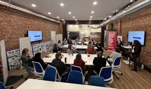 An overview picture from the side of a room filled with people sitting at tables, watching a presentation being given.