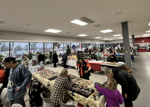 Tables in the cafeteria are filled with vendors at the Indigenous Arts Festival. People are at various tables looking at items.