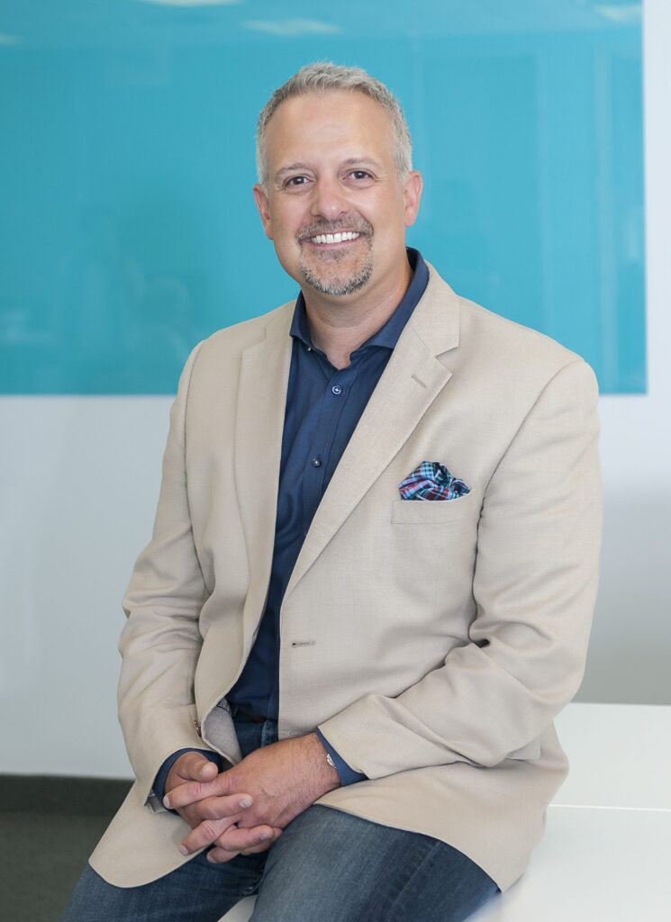 Headshot of Senior Vice-President, External Relations and Business Development, Jeremy Laurin. He's seated on the edge of a table and is wearing a beige suit jacket.