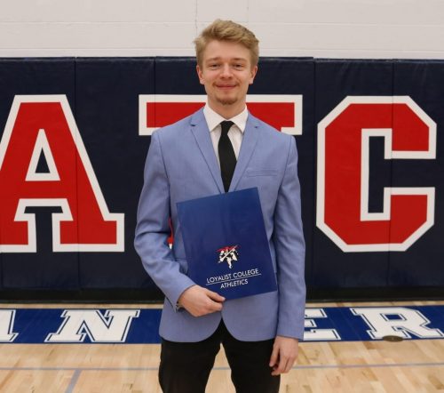 Image of Josh Roberts, a General Arts and Science student, wearing a blue suit jacket, holding a Loyalist Lancers Athletics folder. The background is the wall of the Loyalist gym, with blue padded pats.