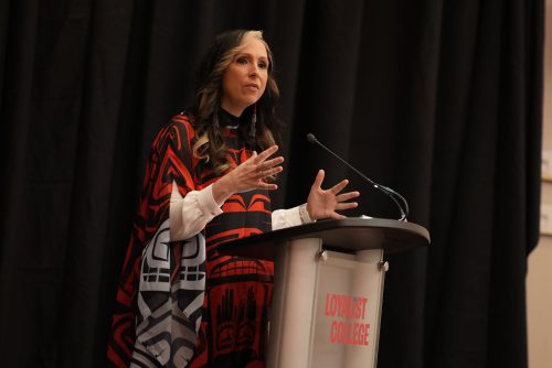 An image of Pam Palmater standing at a Loyalist College podium in front of a black background, talking to an audience not pictured.