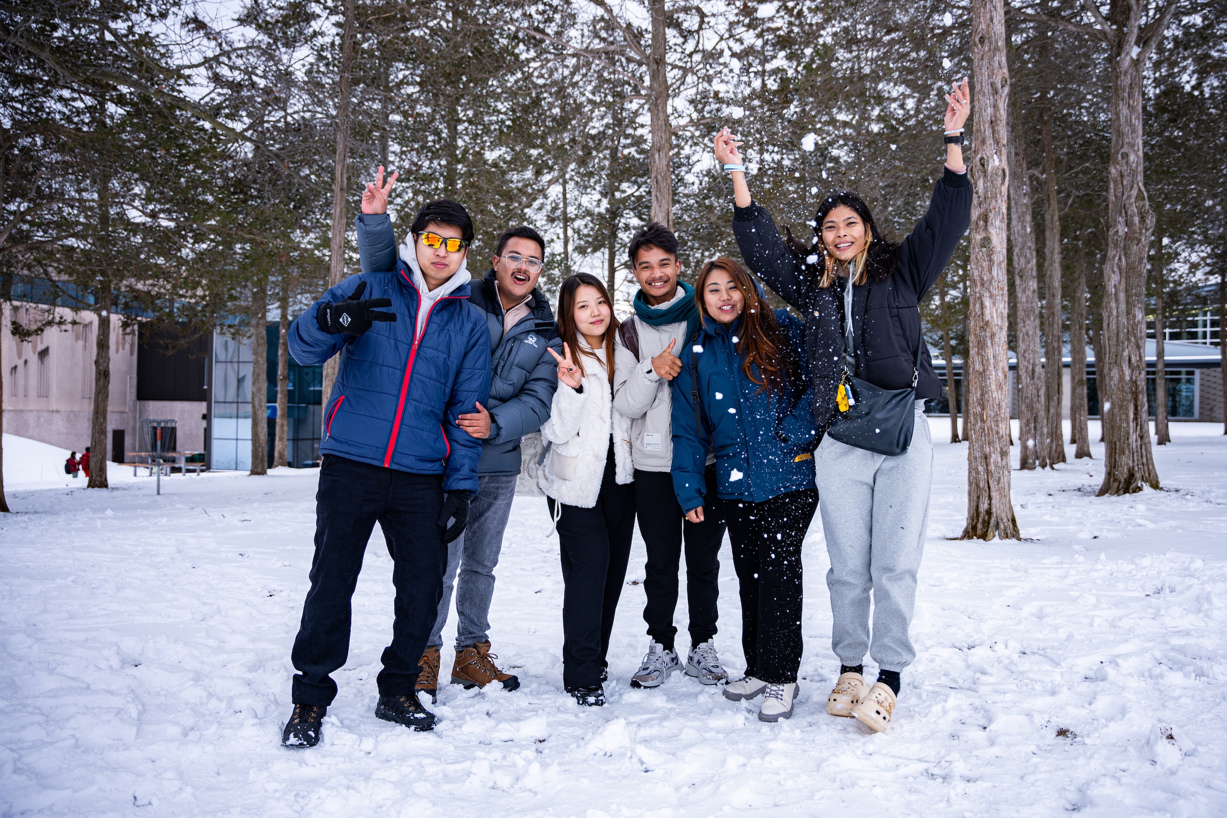 A group of six students standing in the snow, dressed in winter clothing. Some are posing with peace signs and smiles, and one person throws snow into the air as they enjoy the winter scenery.