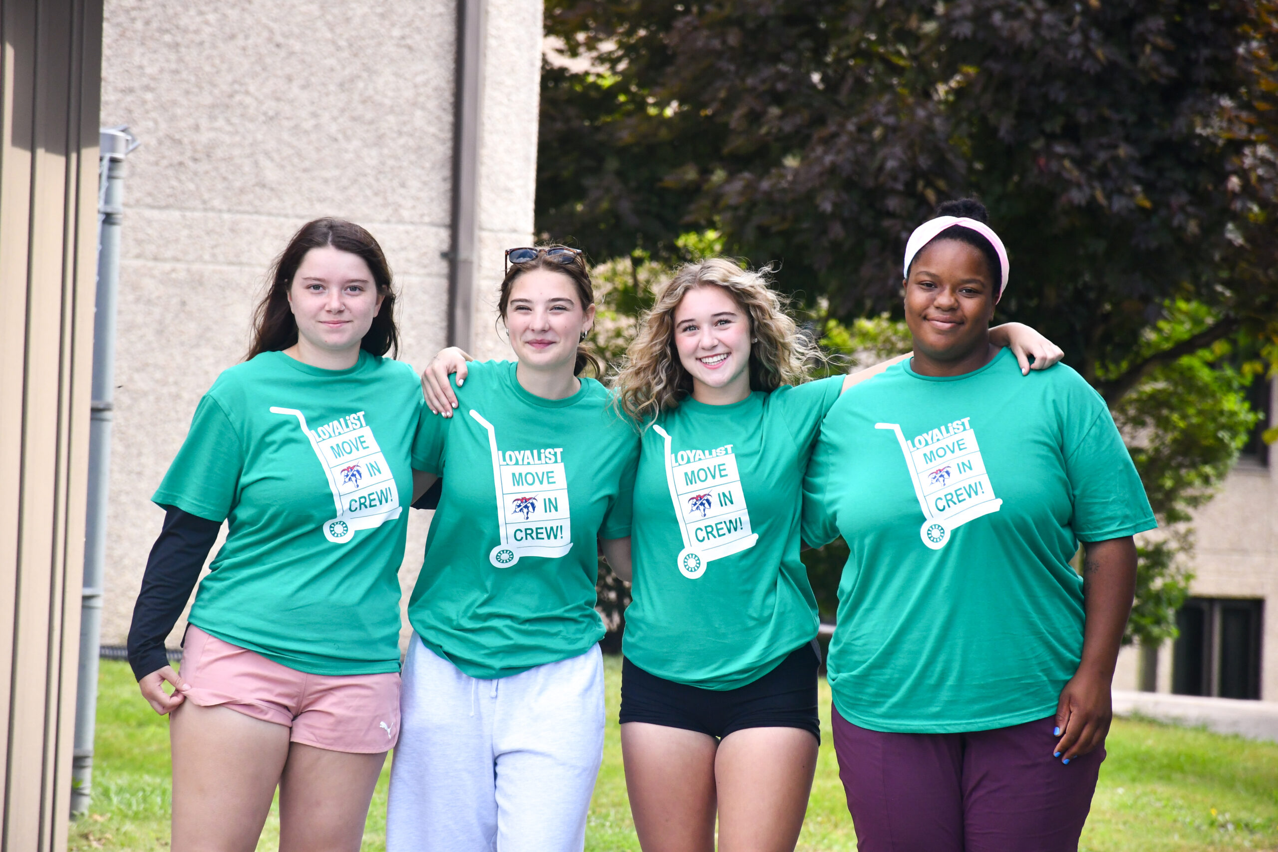 Four young women wearing matching green T-shirts with 'Loyalist Move In Crew!' printed on them. They are standing outdoors, smiling, with their arms around each other.