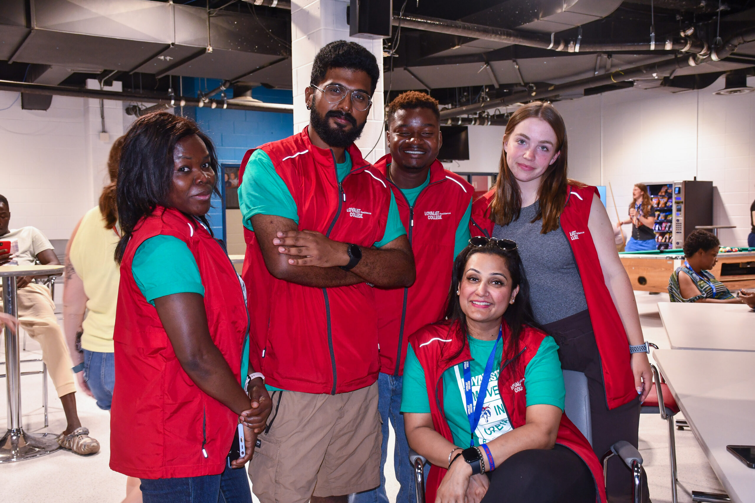 Five students wearing red Loyalist College vests and green shirts standing together in the Shark Tank Pub at the Belleville location, smiling for the camera. One person is seated while the others stand around her.