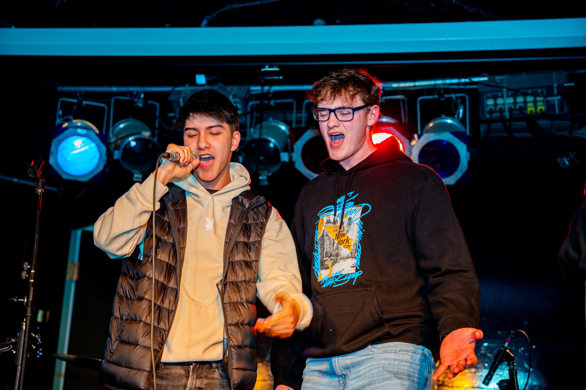 Two young men singing on stage, one holding a microphone, both singing, and both wearing casual clothes. The stage is lit with blue and red spotlights in the background.