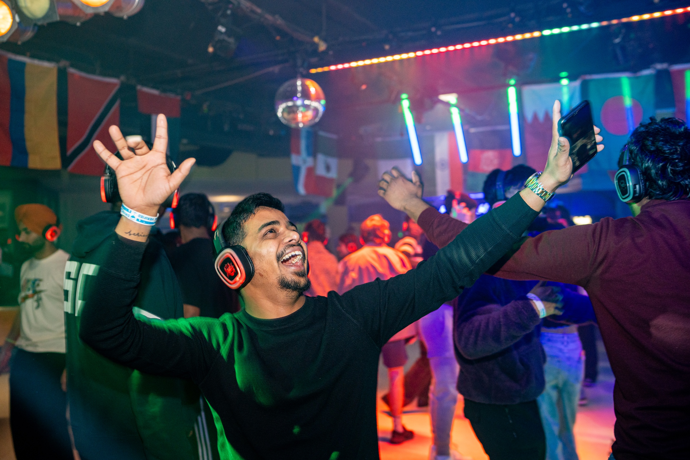 A male student wearing headphones, smiling and dancing with his arms raised during a silent disco event. Colourful lights and international flags hang from the ceiling, with other people dancing in the background.