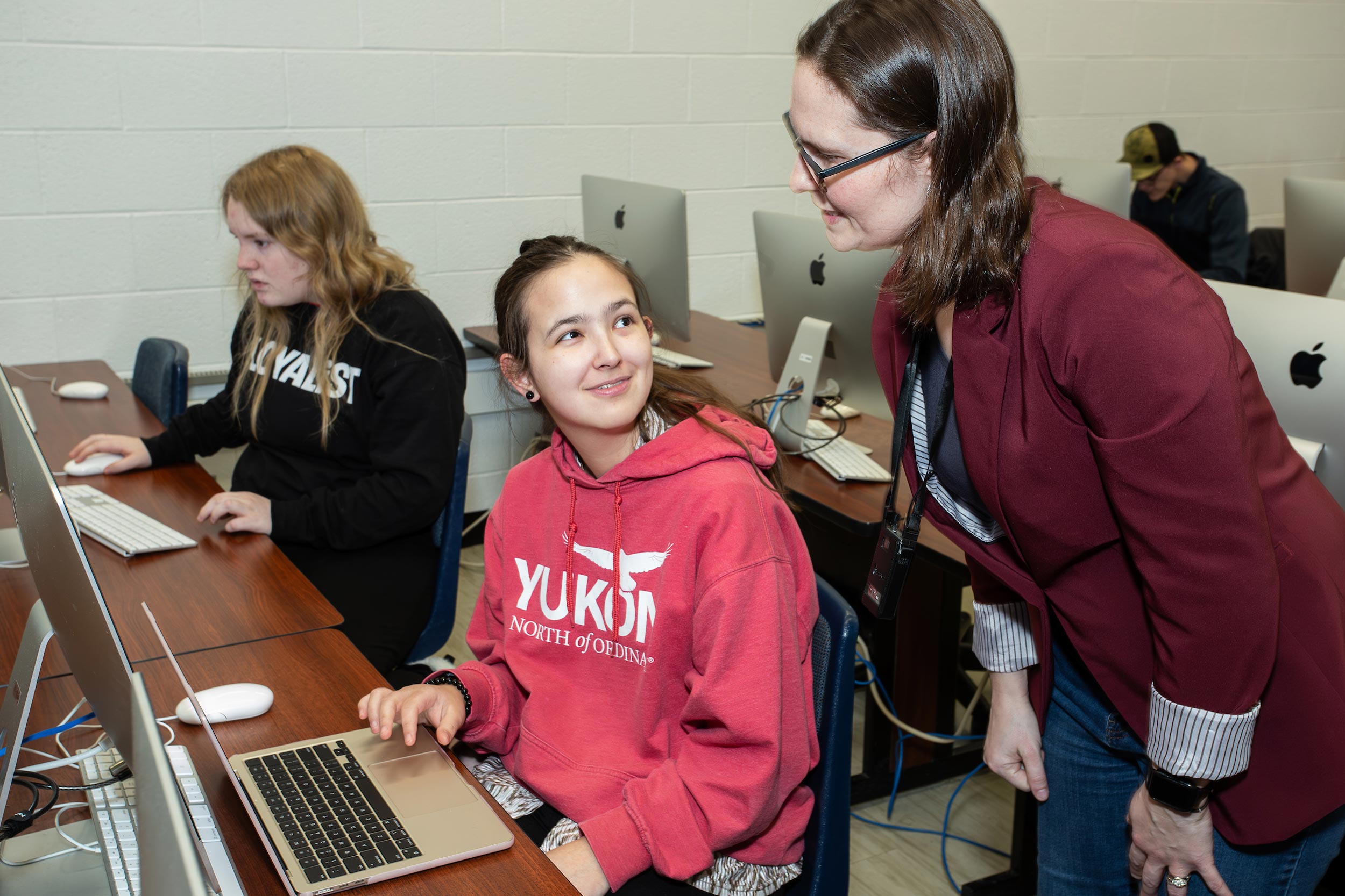 A student in a red Yukon sweatshirt sits at a desk with a laptop, smiling at a woman in a maroon blazer who is leaning over to speak with her. Another student is seated in the background working on a computer in a classroom filled with desktop computers.