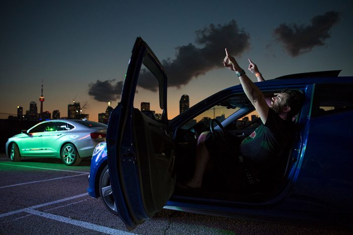 Man sitting in the driver’s seat of a blue car at dusk with the door open, gesturing with both hands pointing upward. A green-lit car is parked nearby, and the city skyline with the CN Tower is visible in the background under a cloudy sky.