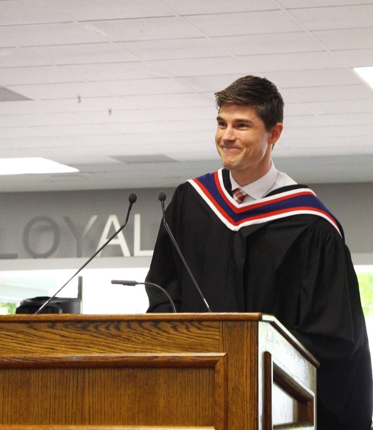 Loyalist College graduate Ryan Snoddon wearing black graduation gown with a sash and hood. He's standing at a podium with an open, well lit space behind him.