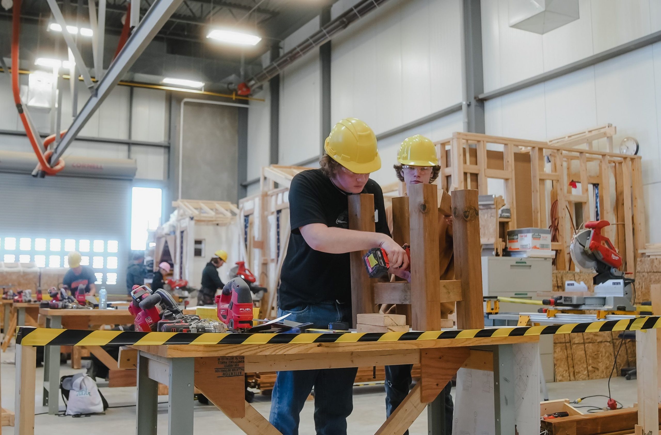 Two students in a workshop, working on constructing an object out of wood. Both wearing yellow hard hats.