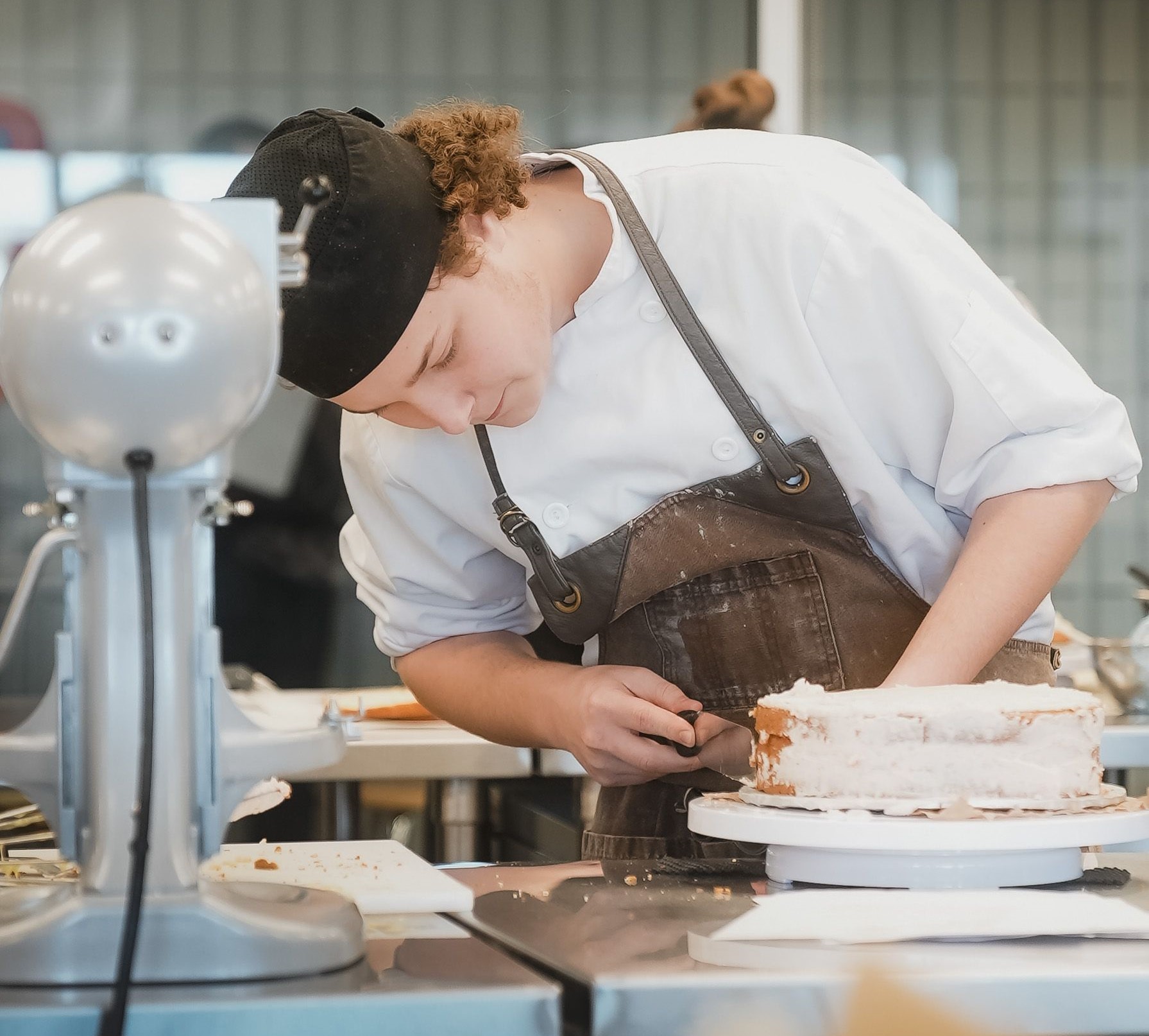 A student in the culinary learning space, wearing an apron and black hat, frosting a cake.