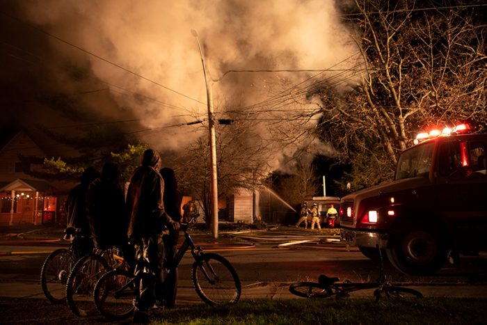 Nighttime scene of a house fire with thick smoke rising into the sky. A group of people, some with bicycles, stand watching from across the street. Firefighters can be seen in the background working to extinguish the flames, while a fire truck with flashing lights is parked nearby.