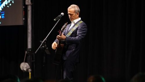 Colonel Chris Hadfield, on a stage in a dark room at a microphone holding a guitar. There's a spotlight on him, otherwise the room is dark.