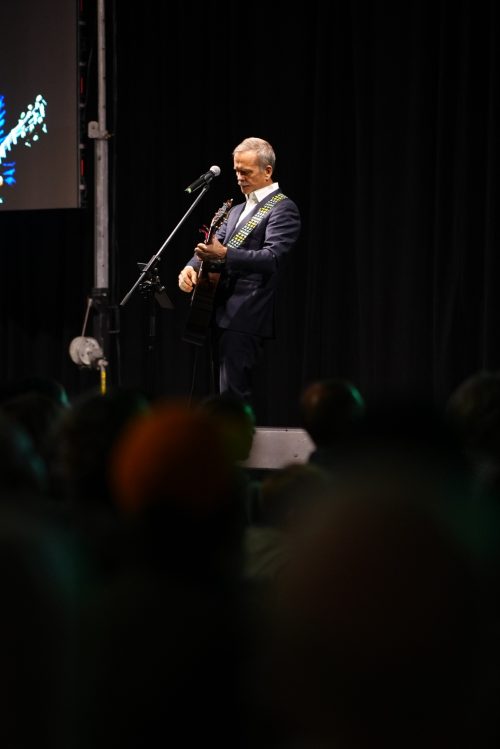 Colonel Chris Hadfield, on a stage in a dark room at a microphone holding a guitar. There's a spotlight on him, otherwise the room is dark.