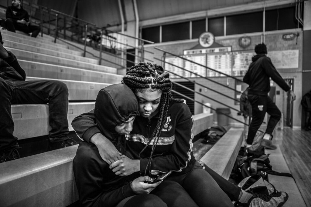 Black and white photo of two people sitting in bleachers. One person is wearing a hoodie with the hood up, while the other, with braided hair, leans in closely and hugs them as they both look at a phone. The setting is a gym with other people and bleachers in the background.