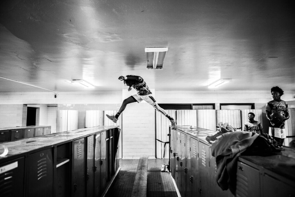 Black and white photo of a person mid-air, jumping over a row of lockers in a locker room. Three other individuals, wearing 'Jags' jerseys, watch from the side. The setting is a dimly lit room with fluorescent lights overhead, and the lockers are lined up along the floor.