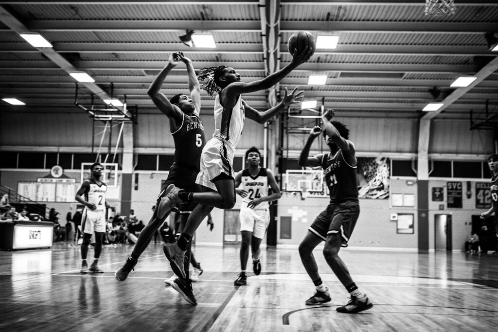 Black and white photo of a basketball game in action. A player in a white jersey is mid-air, reaching toward the hoop with the ball while being defended by two opposing players in dark jerseys. Other players are visible in the background, and the game takes place in an indoor gymnasium.