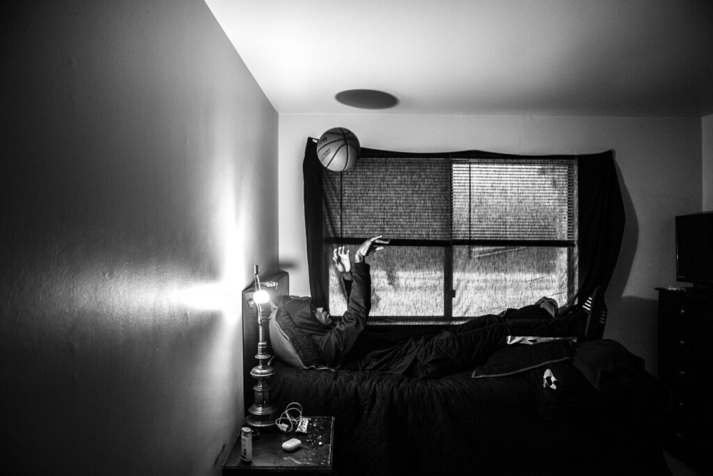 Black and white photo of a person lying on a bed in a dimly lit room, tossing a basketball in the air. The room has a window with partially closed blinds, and the person is positioned under the window, casually throwing the ball upwards. A lamp on the bedside table provides the only source of light.