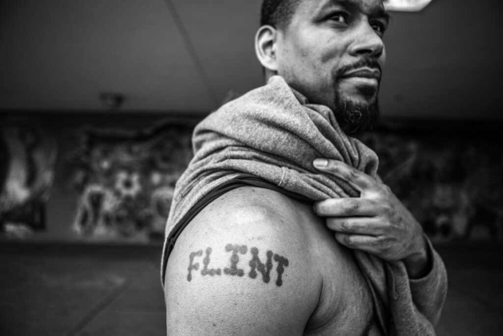 Black and white photo of a man looking off into the distance with the word 'FLINT' tattooed on his upper arm. He is draping a sweater over his shoulder while standing in front of a blurred mural background.